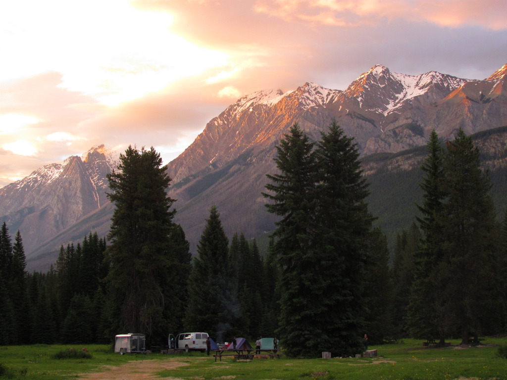 Kampeerplek met tenten en busje in de Canadese Rockies bij zonsondergang, met besneeuwde bergtoppen.