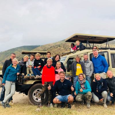 Familiegroep bij safari-jeep tijdens familiereis Tanzania en Zanzibar, met heuvelachtig savannelandschap op de achtergrond.
