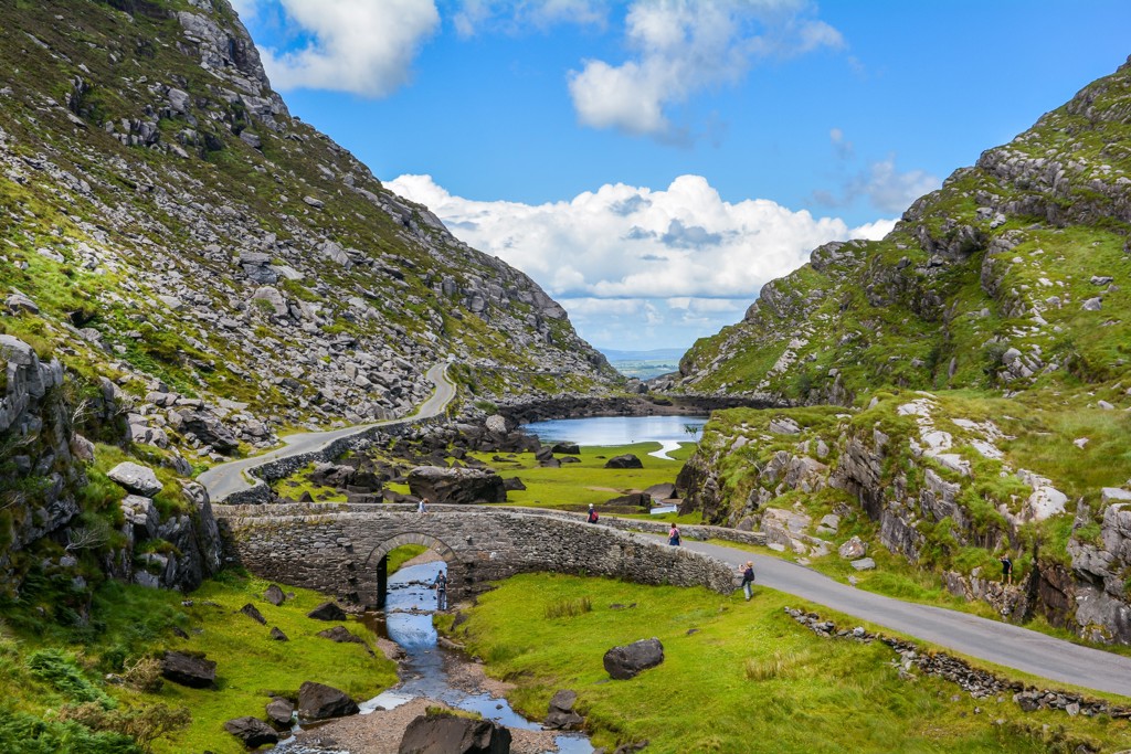 Stone bridge and winding road through the Gap of Dunloe in Ireland, surrounded by green mountains and a lake.