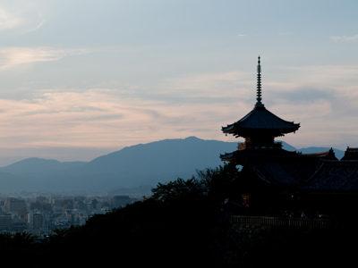 Silhouet van een Japanse tempel bij zonsondergang, met bergen en stad op de achtergrond.