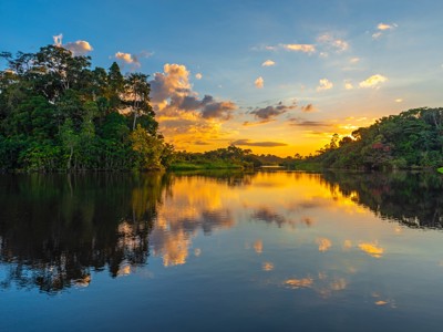Zonsondergang boven een brede rivier in het Amazonegebied van Ecuador, met regenwoud en wolken weerspiegeld in het water.