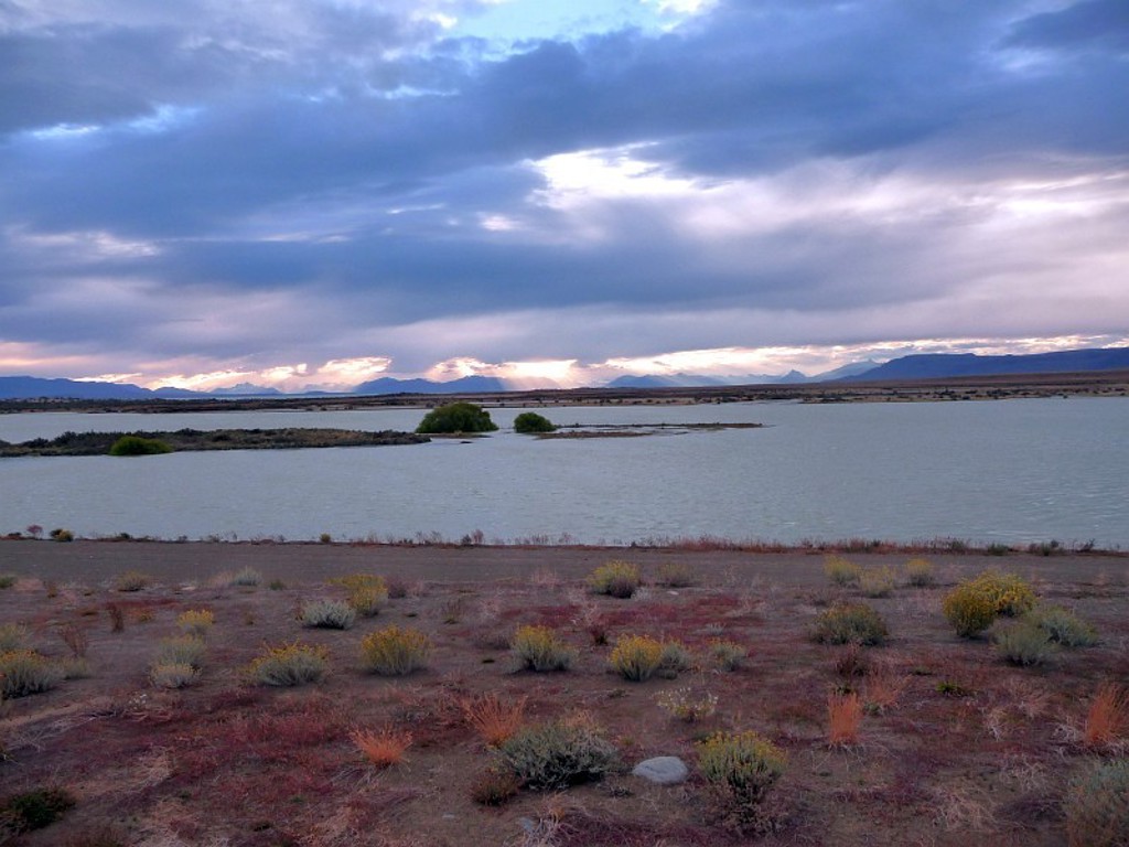 Azuurblauw meer in Patagonië met eilandjes en bergsilhouetten onder een dramatische avondlucht.