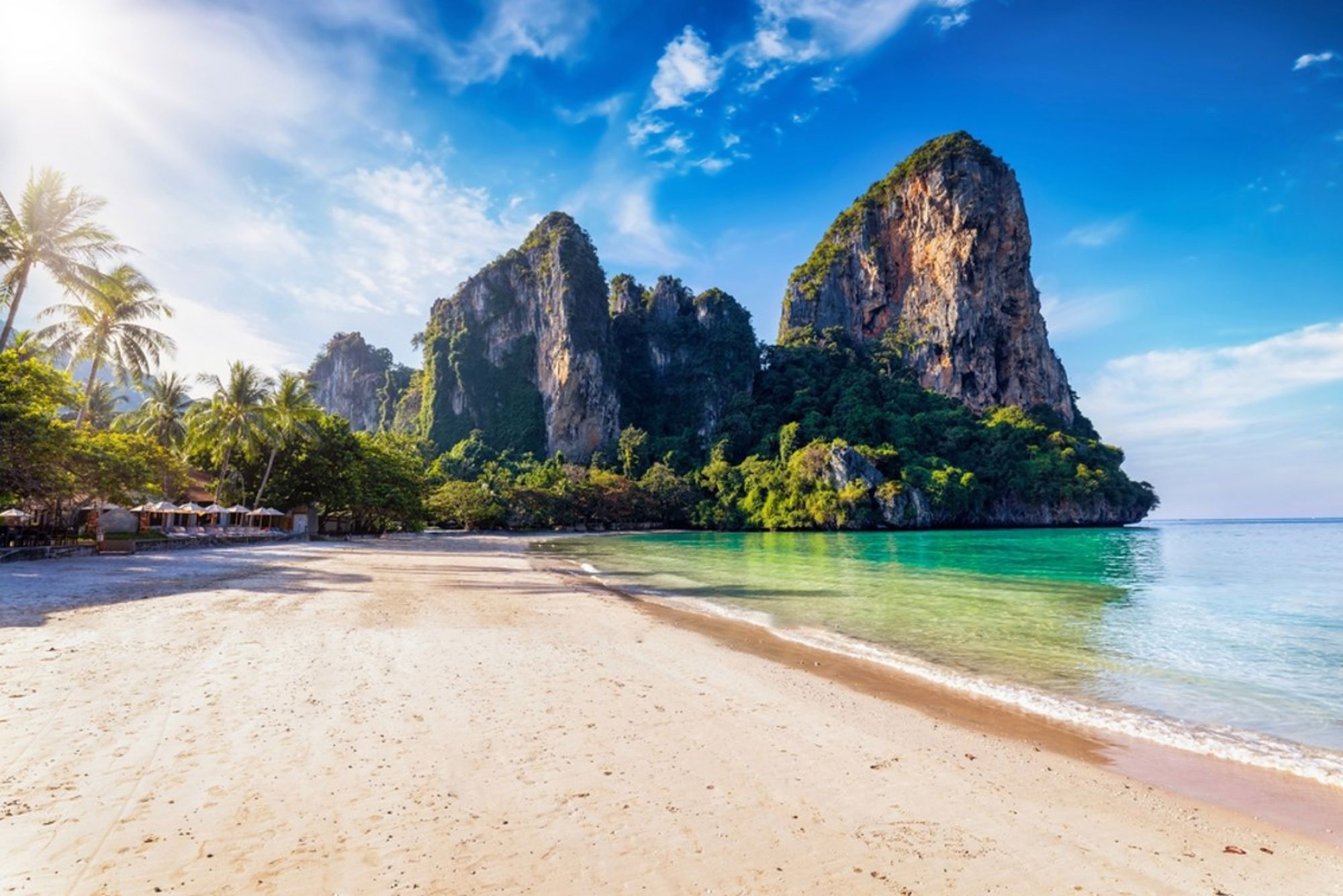 Zandstrand in Thailand met palmbomen, turquoise zee en hoge kalksteenrotsen onder een blauwe lucht.
