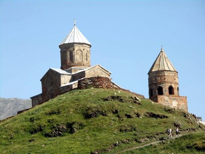 Gergeti Drievuldigheidskerk bij Kazbegi in Georgië, op een groene heuvel met wandelpad en blauwe lucht.