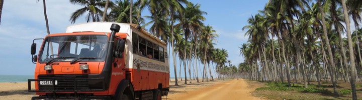 Oranje reisbus op zandweg langs palmbomen aan de kust in Benin.