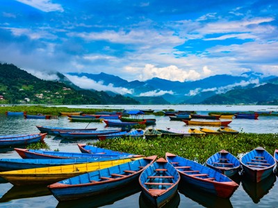 Kleurrijke roeiboten op het Phewa-meer bij Pokhara, met groene heuvels en wolken boven de Himalaya.