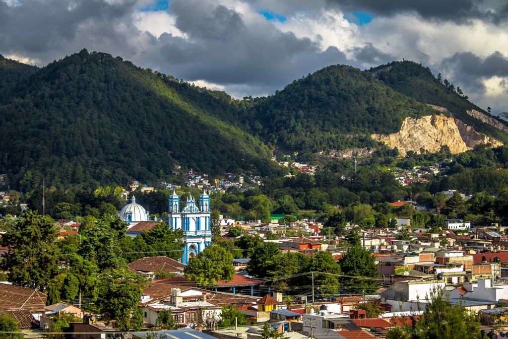 Blauw-witte kerk in een Mexicaans bergdorp, omringd door groene heuvels en dramatische wolkenlucht.