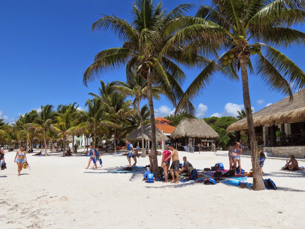 Palmbomen en gezinnen op wit zandstrand in Mexico tijdens familiereis, met strandtenten op de achtergrond.