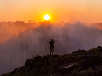 Zonsopkomst boven Victoria Falls met nevel van de watervallen en een reiziger op de rotsen.