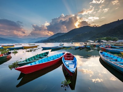 Kleurrijke roeiboten op het Phewa-meer bij Pokhara, met bergen en zonsondergang op de achtergrond.