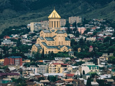 De Heilige Drie-eenheidskathedraal in Tbilisi met de gouden koepel, omringd door de stad en groene heuvels.