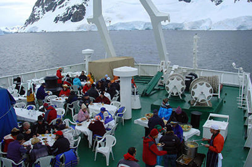 Reizigers eten op het dek van een expeditieschip in Antarctica, met besneeuwde bergen en zee op de achtergrond.
