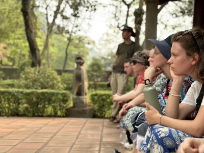 Reisgroep pauzeert in een groene tuin tijdens de Route Vietnam, met een stenen beeld op de achtergrond.