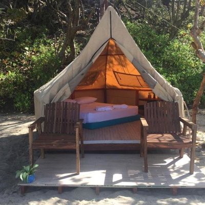 Glampingtent met tweepersoonsbed en houten stoelen op een vlonder, omringd door bomen.