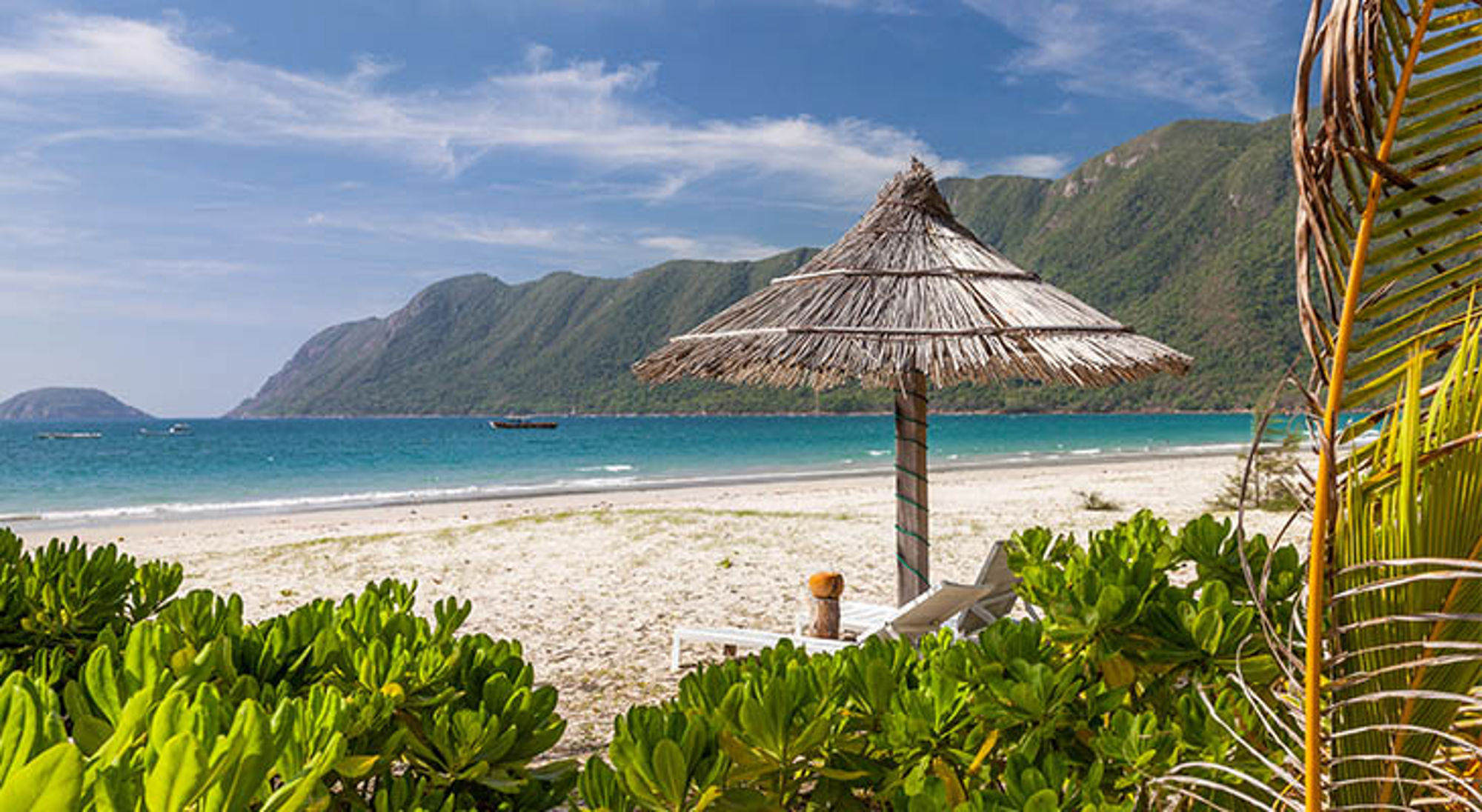 Rieten parasol en ligstoel op zandstrand met turquoise zee en groene bergen op de achtergrond in Vietnam.