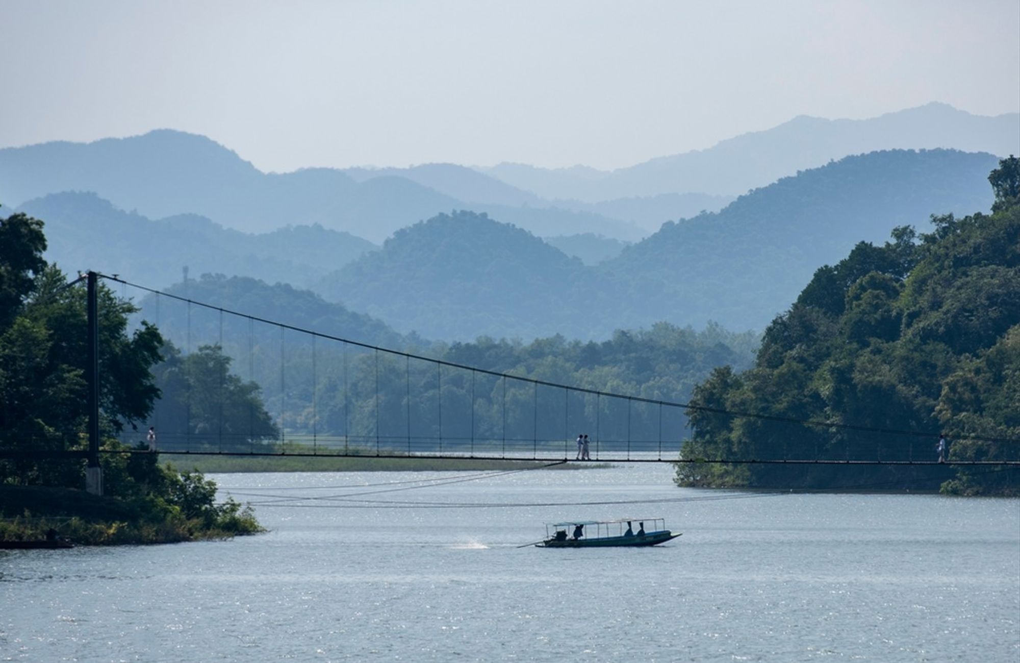 Hangbrug over een rivier met boot, omringd door groene heuvels en bergen in Thailand.