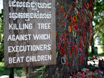 Waarschuwingsbord bij de Killing Tree in Cambodja, met kleurrijke armbandjes aan de boomstam.