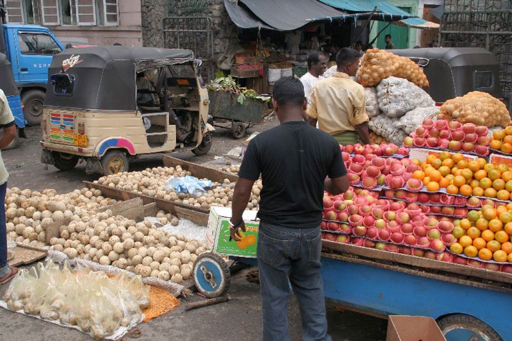 Drukke fruit- en groentemarkt in Sri Lanka met kraam vol appels en sinaasappels en een tuktuk op de achtergrond.