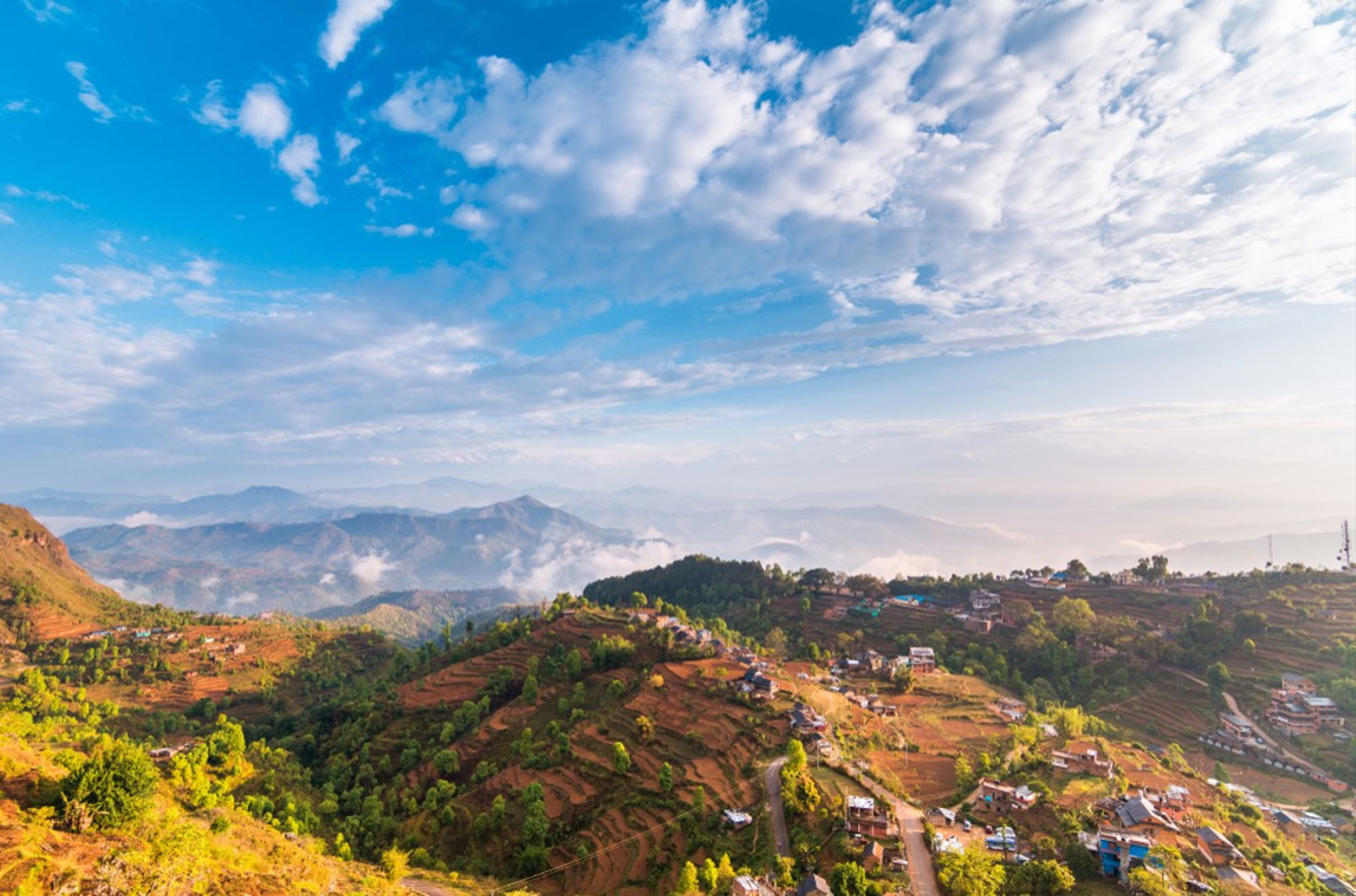 Panorama van heuvels met terrassen en dorpjes in Nepal onder een blauwe lucht met wolken.