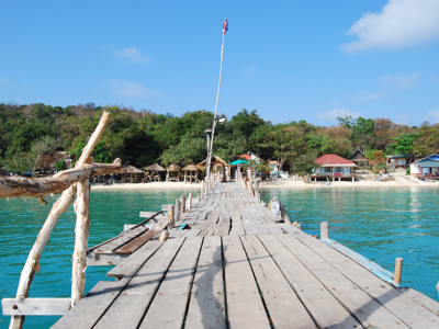Houten pier naar een strand met bungalows aan helderblauw water in Thailand.