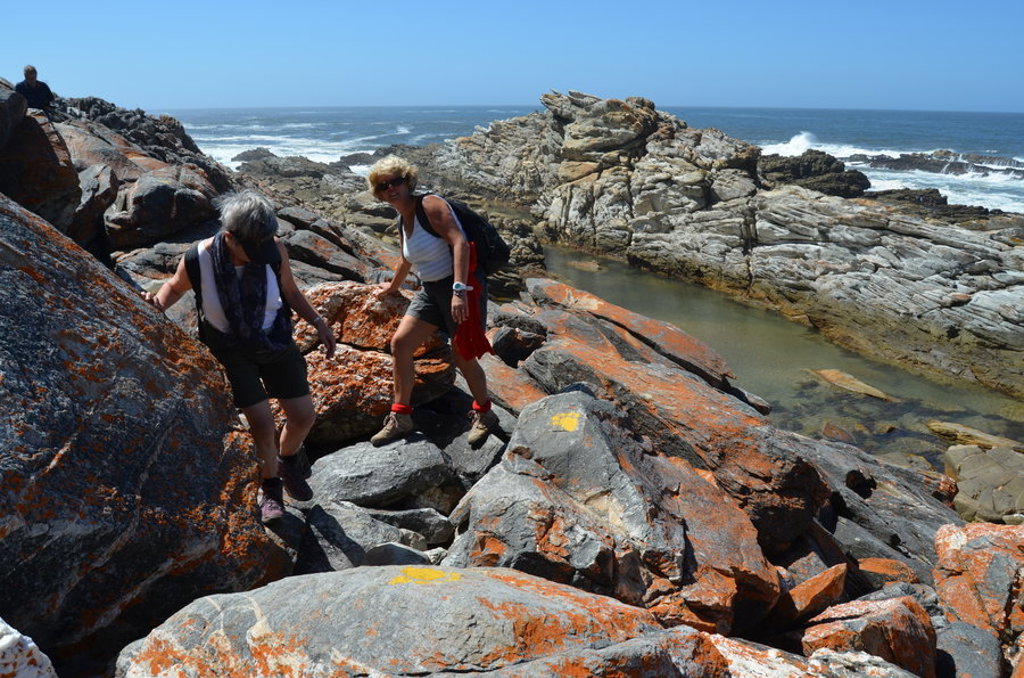 Wandelaars op rotsachtige kustlijn langs de Zuid-Afrikaanse Tuinroute, met uitzicht op zee en kliffen.