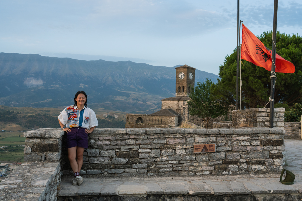 Reiziger bij kasteelmuur met Albanese vlag en klokkentoren, met bergen op de achtergrond in Albanië.