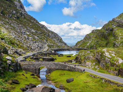 Stenen brug en slingerweg door de Gap of Dunloe in Ierland, met groene heuvels en een meer.