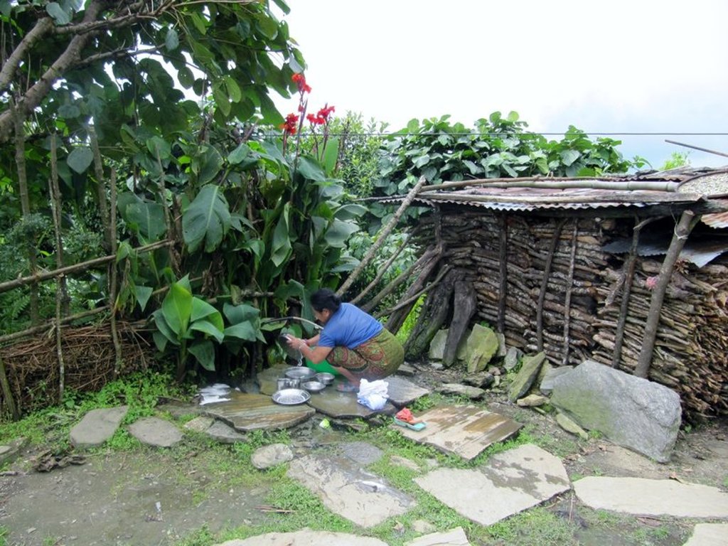 Nepalese vrouw wast pannen bij een waterkraan naast een houten hut in een groen bergdorp.
