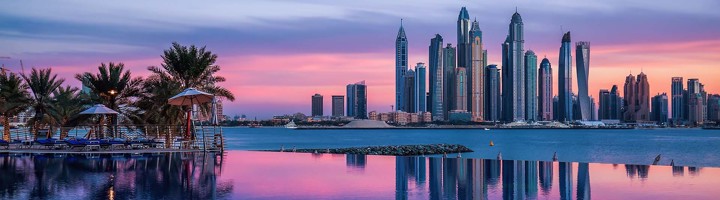 Skyline van Dubai bij zonsondergang, met wolkenkrabbers aan het water en palmbomen op de voorgrond.