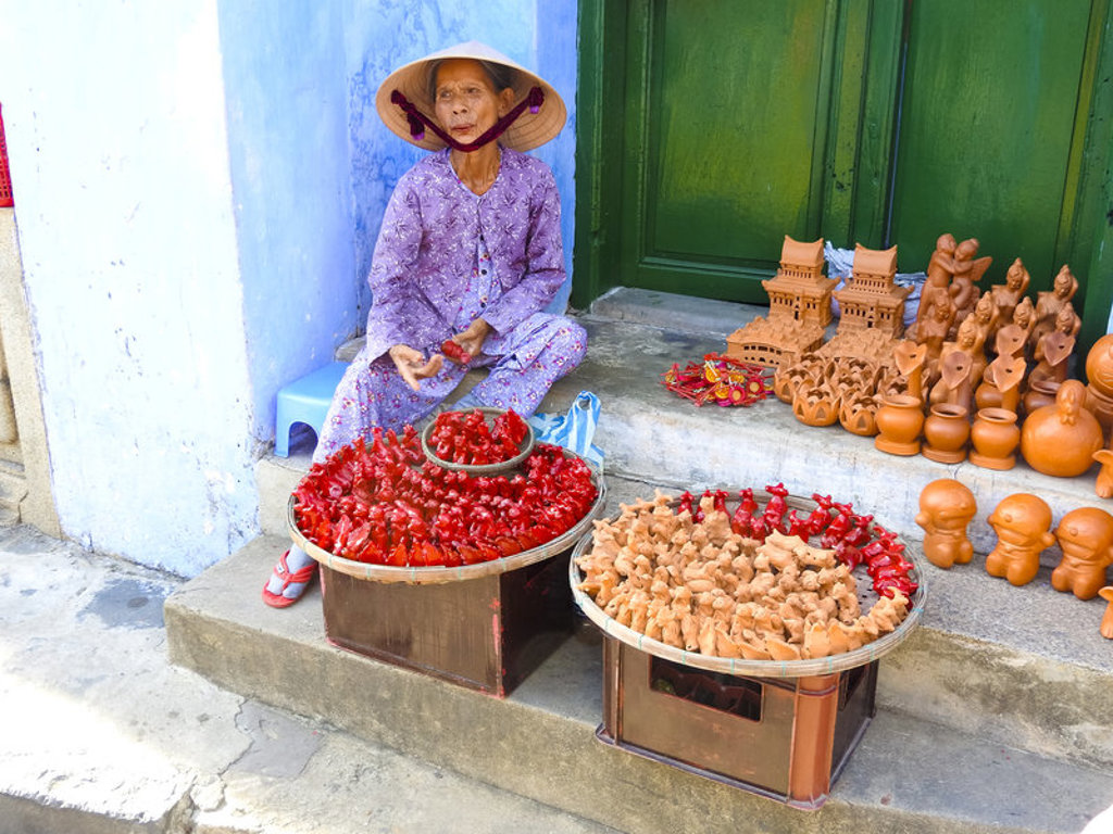 Vietnamese straatverkoper met conische hoed verkoopt rood en beige speelgoed en aardewerk tijdens fietsvakantie Vietnam.