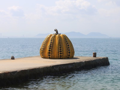 Gele pompoensculptuur met zwarte stippen op een pier aan zee in Japan.
