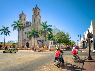 Kathedraal met palmbomen aan een plein in Valladolid, Mexico, met scooters en verkeer op straat.