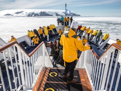 Reizigers op het dek van een expeditieboot in het zee-ijs bij Groenland, met besneeuwde bergen op de achtergrond.