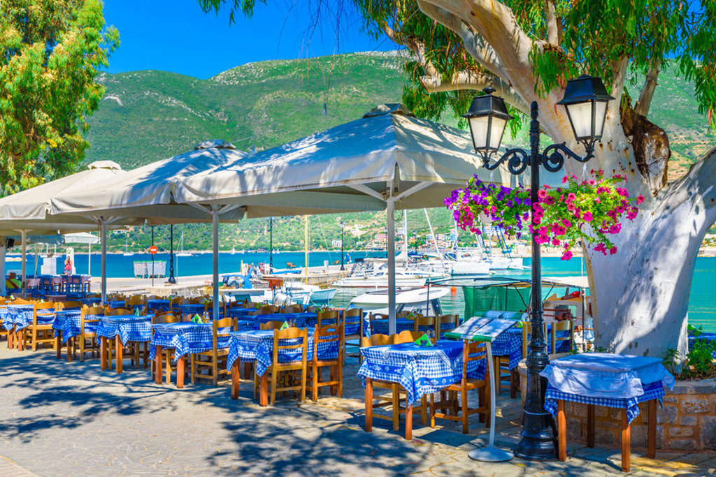 Terras aan de haven in Griekenland met tafels onder parasols en uitzicht op zeilboten en groene bergen.