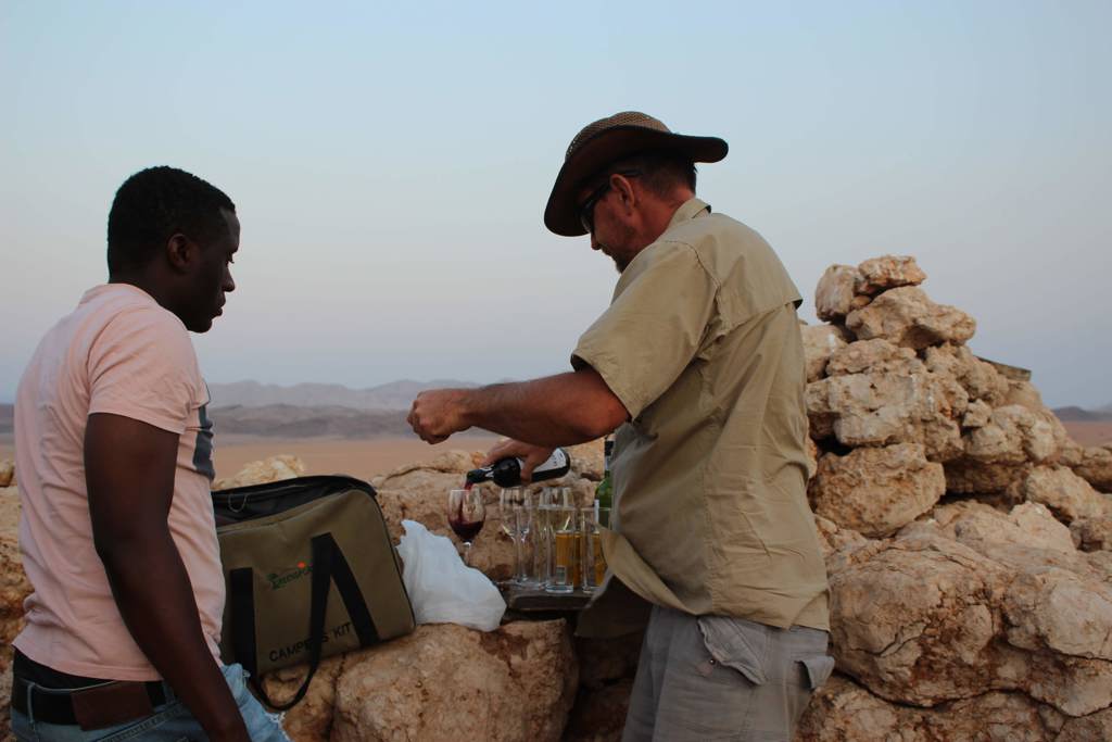 Sundowner in Namibië: gids schenkt wijn in bij rotsen met uitzicht op de woestijn.