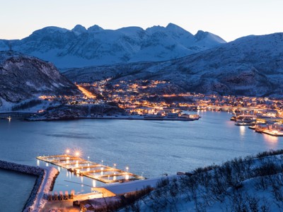 Verlicht kustplaatsje aan een fjord in Noord-Noorwegen, omringd door besneeuwde bergen bij schemering.