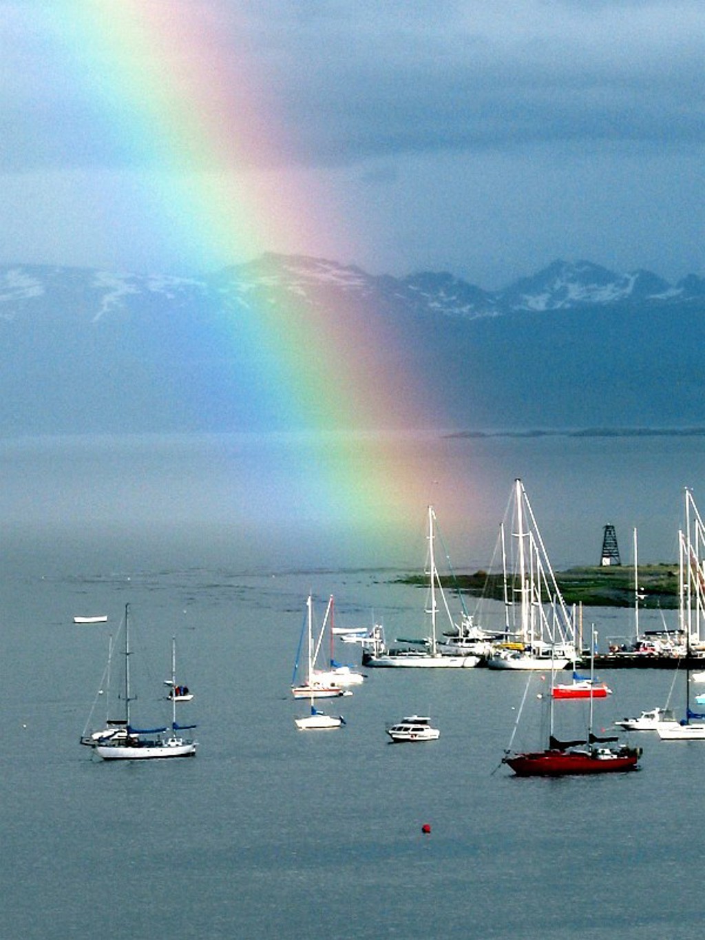 Regenboog boven een Patagonisch meer met zeilboten en besneeuwde bergtoppen op de achtergrond.