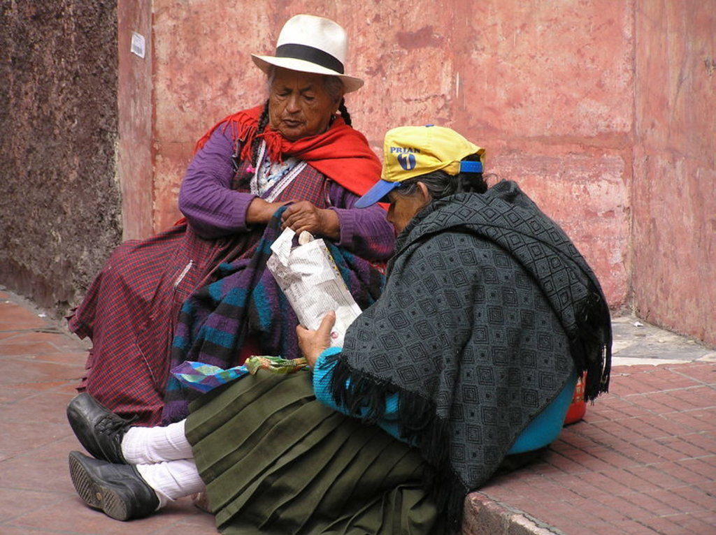 Twee vrouwen in traditionele kleding zitten op straat in Ecuador en lezen samen een krant.