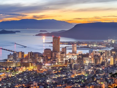 Avondzicht op de skyline van Kagoshima aan zee, met verlichte gebouwen en bergen bij zonsondergang in Japan.