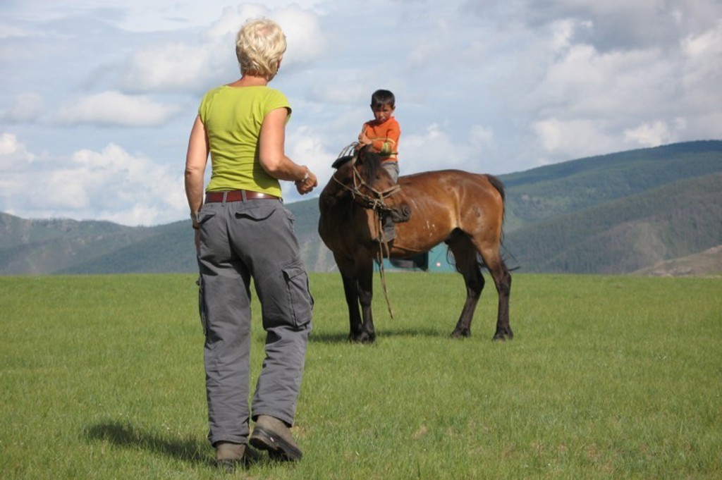 Reiziger op de Mongoolse steppe kijkt naar een kind op een paard, met bergen en blauwe lucht op de achtergrond.