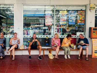 Reizigers pauzeren bij een 7‑Eleven in Thailand tijdens de Route Thailand 21 dagen, met snacks en drankjes op de bank.