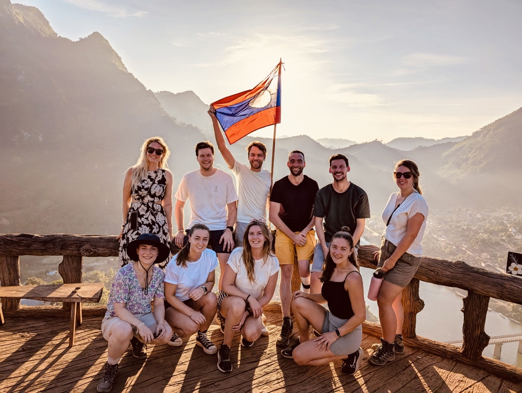Groepsfoto van 22-35ers reisgenoten met Laos-vlag bij uitzichtpunt in de bergen van Laos.