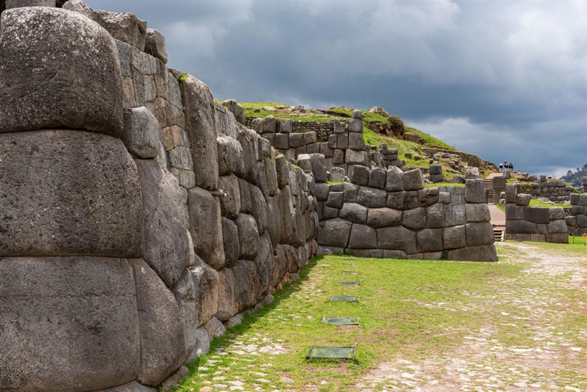 Inca-stenen muren van Sacsayhuamán bij Cusco, Peru, met grasveld en dreigende wolkenlucht.