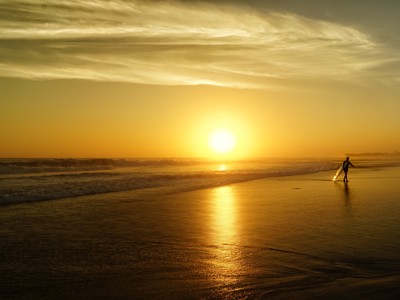 Zonsondergang op het strand van Bali met een surfer die zijn surfplank draagt langs de branding.