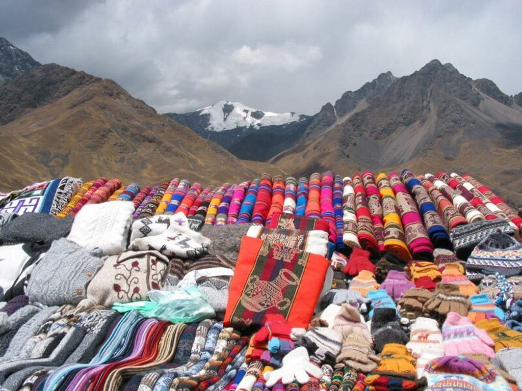 Kleurrijke handgemaakte textiel en souvenirs op een bergmarkt in Peru, met besneeuwde Andes op de achtergrond.