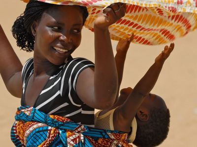 Glimlachende vrouw met kind op de rug, beschut door een kleurrijke doek tijdens de Voodoo reis  in Ghana, Togo en Benin.