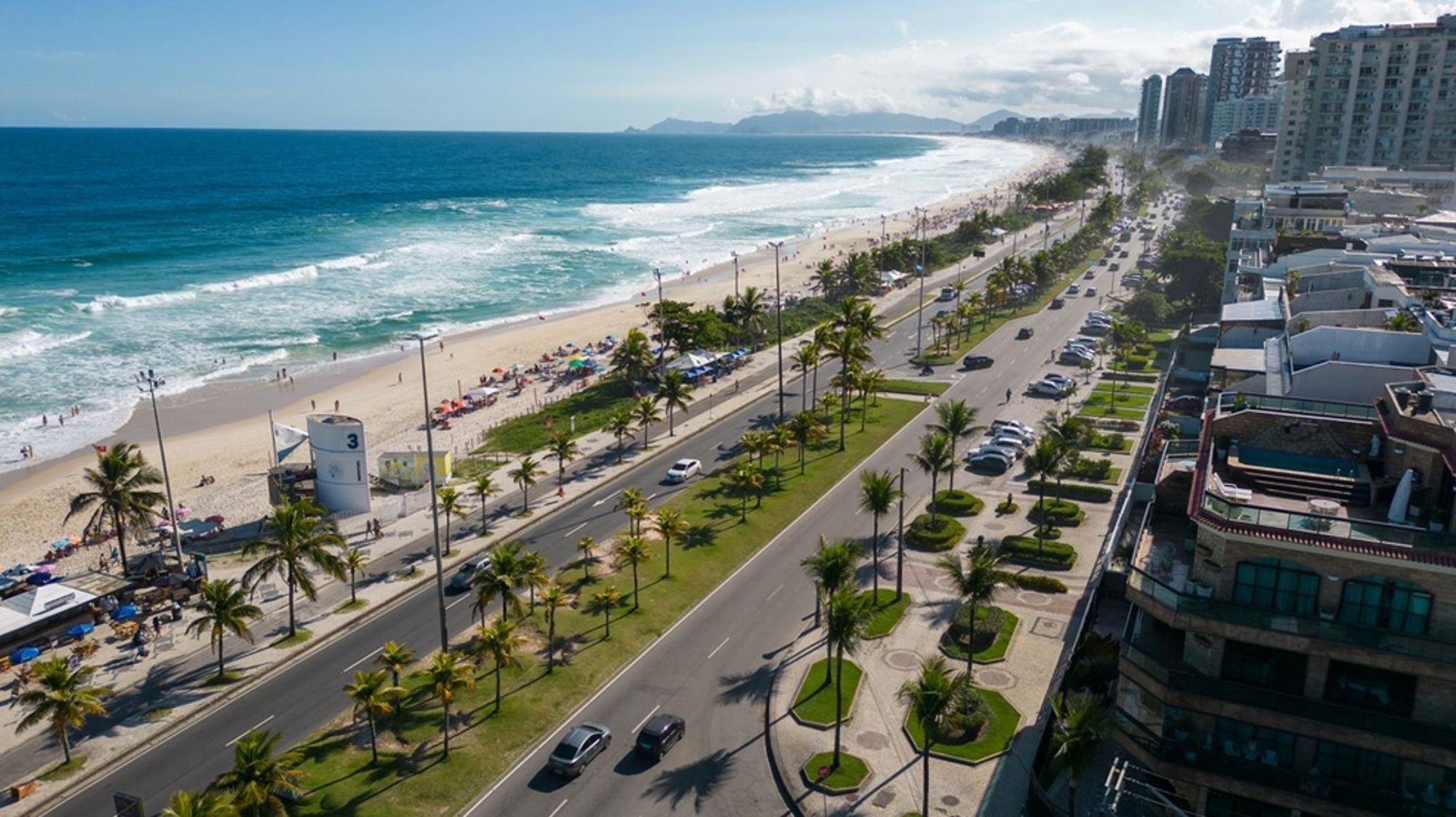 Panoramisch uitzicht op Barra da Tijuca-strand in Rio de Janeiro, met boulevard, palmbomen en hoogbouw.