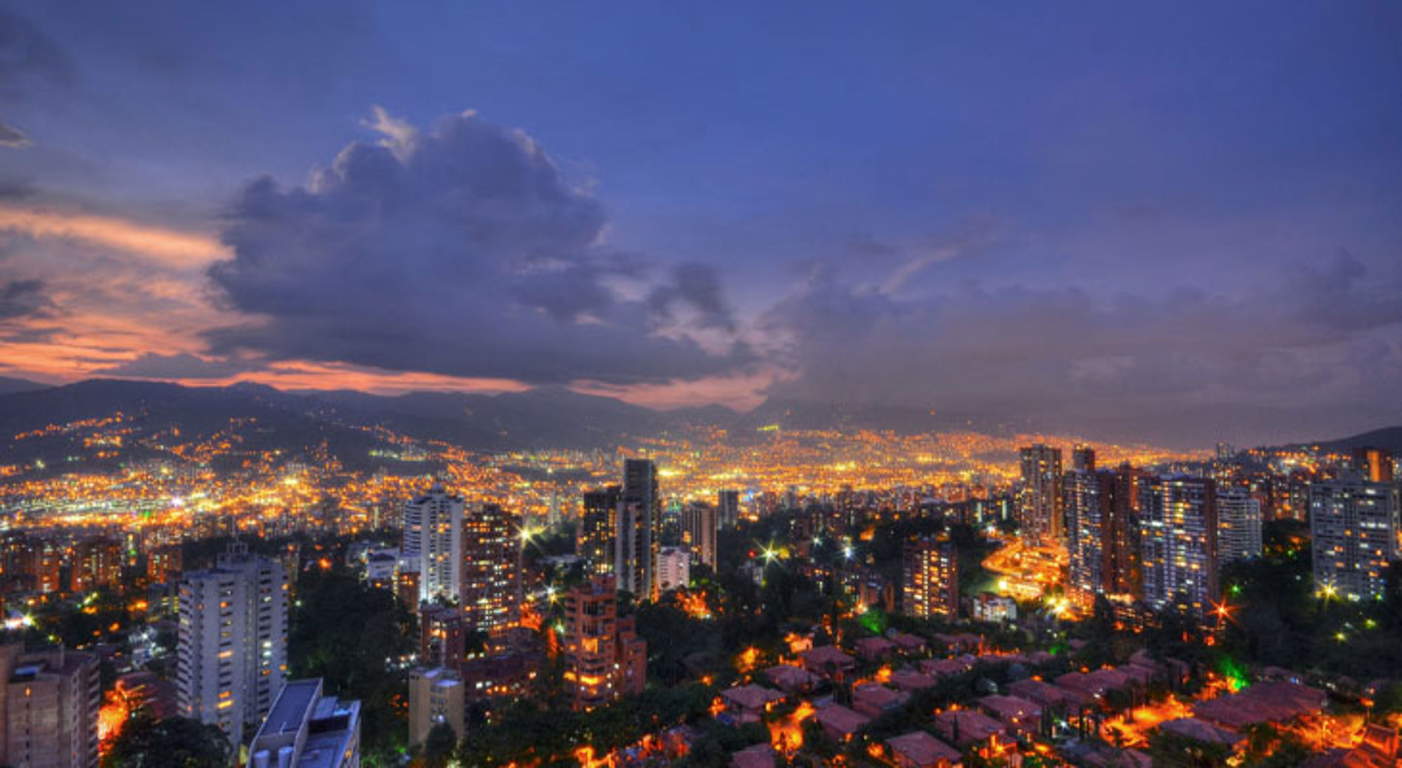 Avondpanorama van Medellín, Colombia, met verlichte skyline en bergen onder een dramatische wolkenlucht.