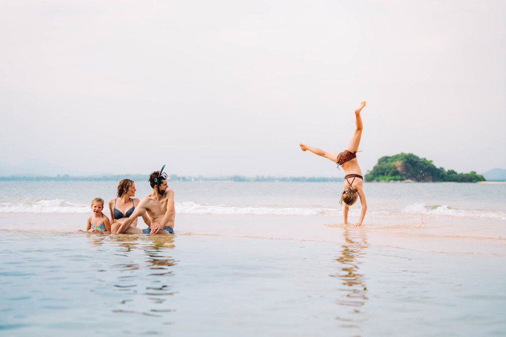 Gezin ontspant op het strand in Thailand, terwijl een kind een handstand doet bij de branding.