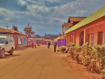 Stoffige straat in een dorp in Tanzania met kleurrijke gebouwen en wandelaars onder een bewolkte lucht.